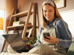 Young woman on her laptop looking at her phone smiling.
